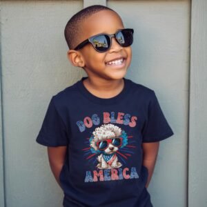 a boy wearing a navy tee featuring a graphic of a cartoon style poodle wearing an american flag bandana with the words dog bless america in red and blue