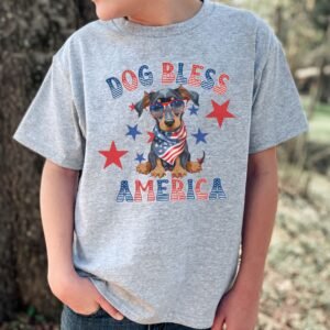 a boy wearing a sport grey tee featuring a graphic of a cartoon style doberman wearing an american flag bandana with the words dog bless america in red and blue