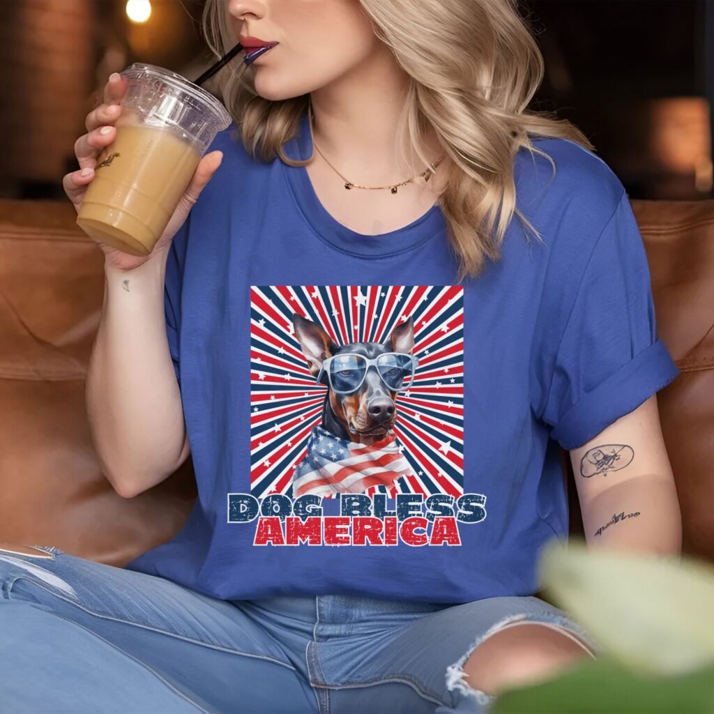 a woman sitting in a coffee shop wearing a heather blue tee with a patriotic background and an image of a doberman in an american flag bandana and sunglasses that says dog bless america