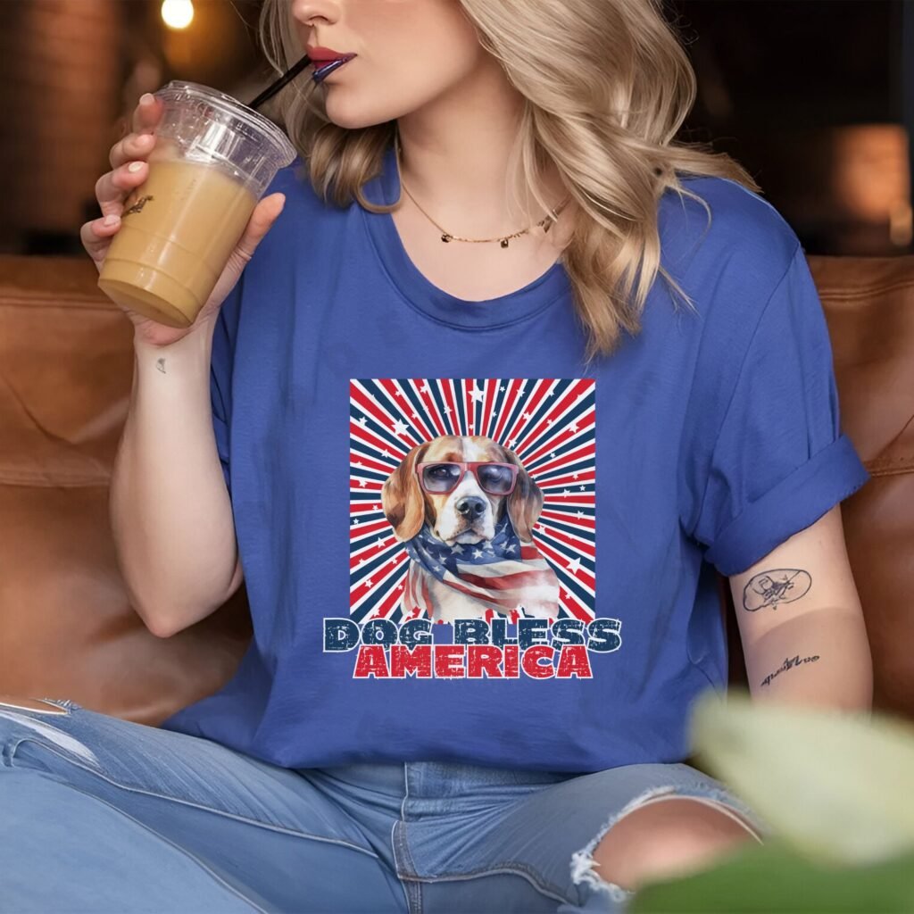 a woman sitting in a coffee shop wearing a heather blue tee with a patriotic background and an image of a beagle in an american flag bandana and sunglasses that says dog bless america