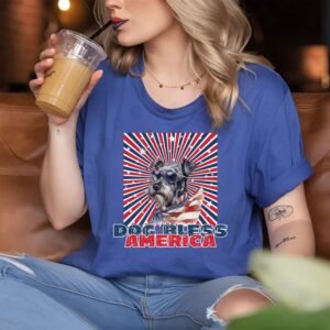 a woman sitting in a coffee shop wearing a heather blue tee with a patriotic background and an image of a schnauzer in an american flag bandana and sunglasses that says dog bless america