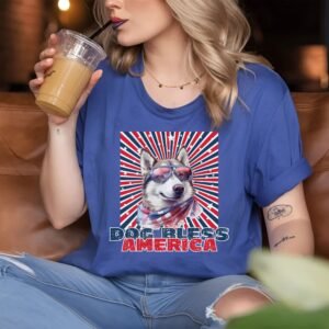 a woman sitting in a coffee shop wearing a heather blue tee with a patriotic background and an image of a husky in an american flag bandana and sunglasses that says dog bless america
